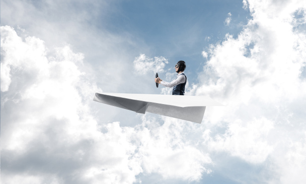 Geschäftsmann mit Fliegermütze sitzt in Papierflieger und hält Lenkrad. Pilot steuert Papierflugzeug in wolkenlosem blauem Himmel. Extreme Luftfahrt Hobby. Himmelspanorama mit flauschigen Wolken. - Foto, Bild