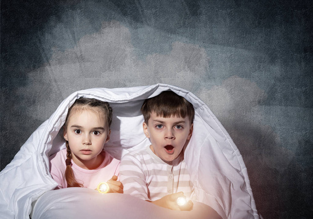 Scared children with flashlights lying in bed. Little sister and brother hiding under blanket together. Covered kids afraid of dark at night on background of grey wall. Night terrors of child. - Фото, зображення
