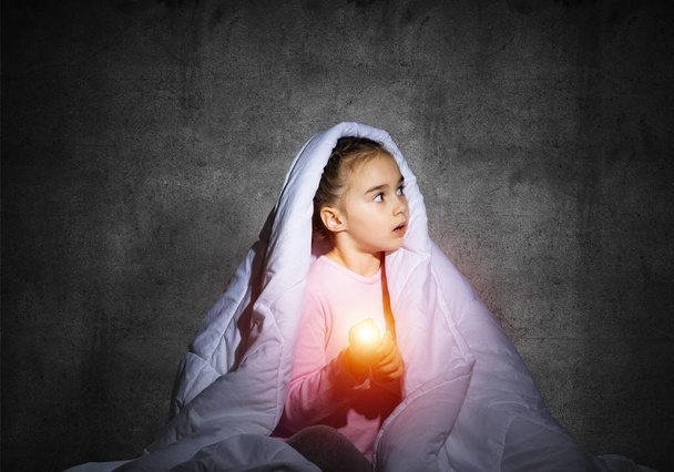 Frightened girl with flashlight hiding under blanket. Scared kid lying in his bed at home. Night terrors of child. Little girl can not sleep at night. Child in pajamas on background of grey wall. - Фото, изображение