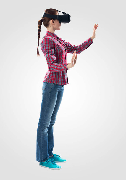 Young woman wearing VR goggles and gesturing in virtual reality. Woman standing and touching air during using VR headset. Studio photo by side view girl against gray background. Cyber technology - Fotoğraf, Görsel
