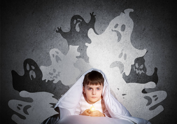 Little child with flashlight hiding under blanket. Halloween scary ghostly monsters on wall. Adorable kid lying in bed at home. Young boy can not sleep at night. Portrait of cute boy in pajamas. - Photo, Image