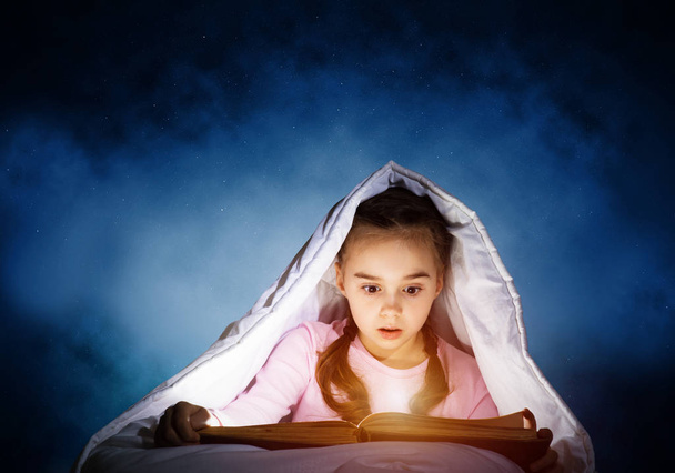 Engrossed little girl reading book in bed before going to sleep. Pretty kid with flashlight hiding under blanket. Beautiful girl in pajamas on deep blue sky background. Child reading fairy tales. - Фото, изображение