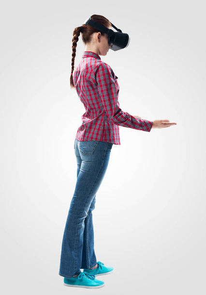 Young woman wearing VR headset and gesturing in air. Woman keep your hands in front of you as something holding. Interacts with cyberspace using gestures. Studio photo by girl against gray background - Photo, Image