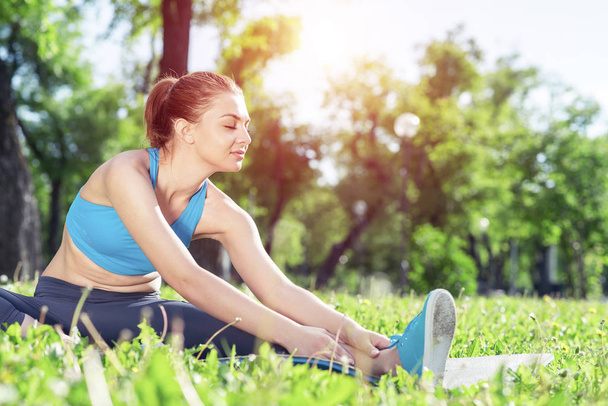 Attractive girl in sportswear doing yoga in park. Young woman sitting in yoga pose on green grass. Sport training outdoor at sunny summer day. Morning stretching exercises and healthy lifestyle. - Foto, Bild