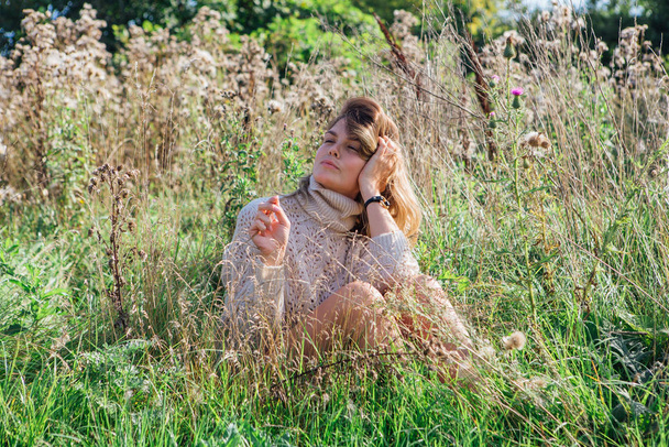 Happy young beautiful woman in sweater and underpants sitting with bare foot in a field of dry agrimony - Фото, зображення