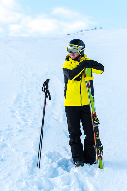 χαρούμενος αθλητής με κράνος στέκεται κοντά σε μπαστούνια του σκι στο χιόνι - Φωτογραφία, εικόνα