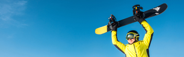 panoramic shot of happy sportsman in helmet holding snowboard above head - Photo, Image