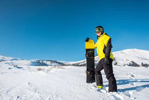 deportista feliz en casco celebración de snowboard y de pie sobre nieve blanca en las montañas
 - Foto, Imagen