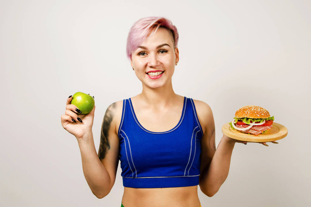 A young beautiful girl smiles and holds hamburger and green apple on a light background. Conception healthy eating and fast unhealthy food - choice problem. - Photo, image