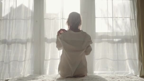 Back view of young Caucasian girl sitting on the bed in the morning. Blond charming woman waking up in her room. Leisure, silence, beauty. - Felvétel, videó