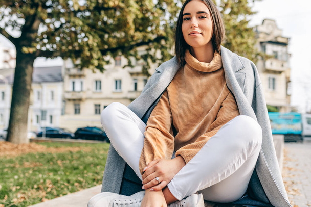 Horizontal outdoor image of beautiful young brunette woman wearing beige sweatshirt, smiling and resting in the city street. Pretty female student relaxing in the city street.  - Valokuva, kuva