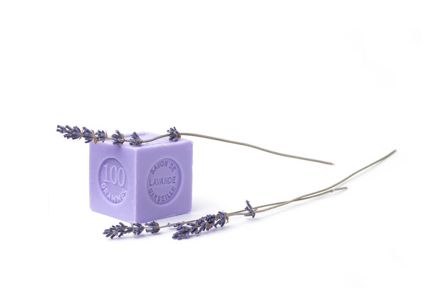 Sabão de lavanda natural de uma forma cúbica de cor lilás com a inscrição em francês: sabão de lavanda de Marselha, 100 gramas e flores de lavanda em um contexto branco
 - Foto, Imagem