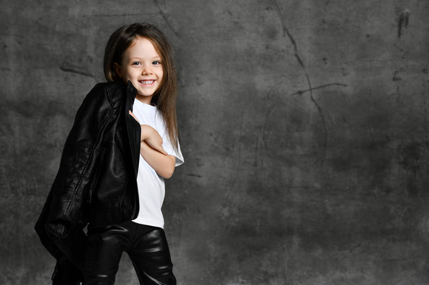 Small smiling cute girl in black and white rock star style casual clothing and white sneakers standing over grey concrete background - Fotografie, Obrázek