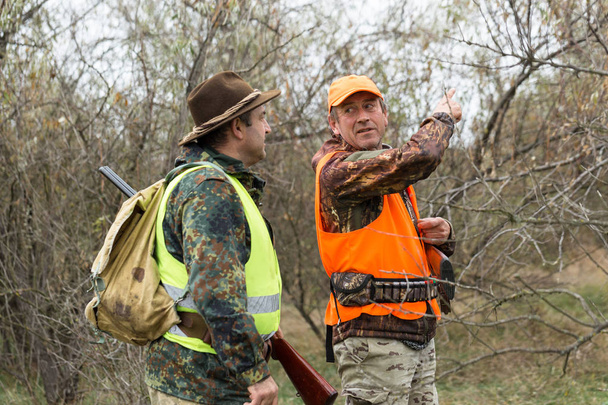 A mans with a gun in his hands and an orange vest on a pheasant hunt in a wooded area in cloudy weather. Hunters with dogs in search of game. - Photo, Image