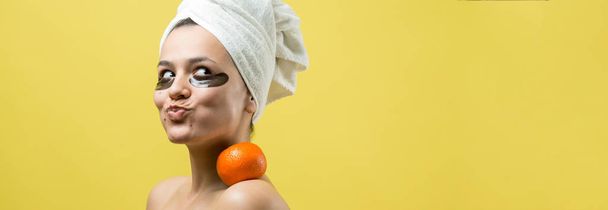 Retrato de beleza da mulher em toalha branca na cabeça com máscara nutritiva dourada no rosto. Skincare limpeza eco orgânico spa cosmético relaxar conceito. Uma menina fica de costas segurando um mandarim laranja
. - Foto, Imagem
