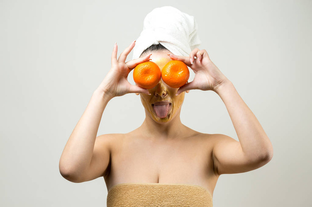 Schönheitsporträt einer Frau in weißem Handtuch auf dem Kopf mit goldener Pflegemaske im Gesicht. Reinigung der Haut Öko-Kosmetik Wellness-Entspannungskonzept. Ein Mädchen steht mit dem Rücken und hält eine orange Mandarine. - Foto, Bild
