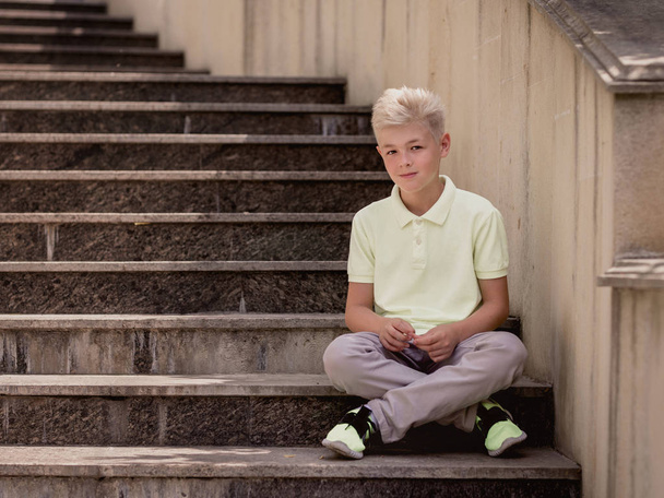 portret van leuke knappe blonde elf oude jongen (tiener) zitten op de trap buiten. Opgroeien, kindertijd, scheiden van concept - Foto, afbeelding