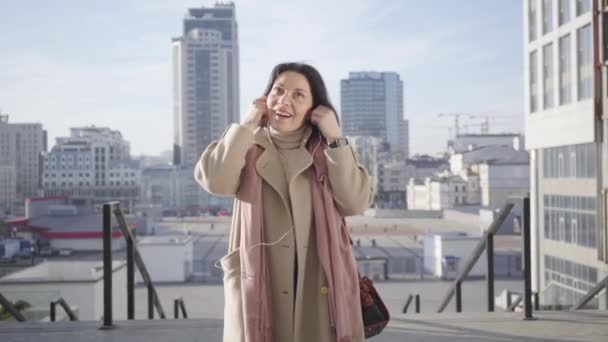 Adult Caucasian brunette woman putting on headphones and enjoying music. Young elegant lady in beige coat resting on city. street Leisure, happiness, lifestyle, hobby. - Filmati, video