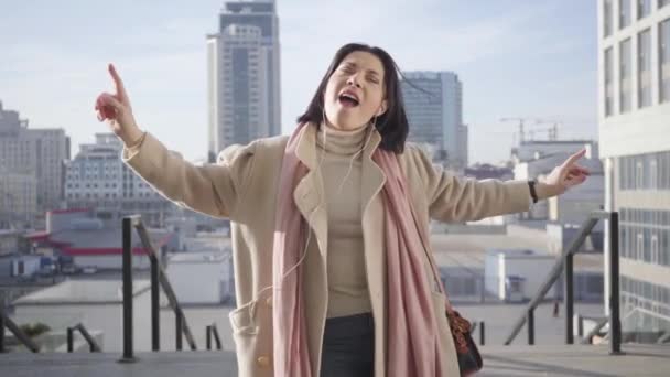 Portrait of cheerful adult Caucasian woman in headphones dancing and singing on city street. Positive brunette businesslady having break. Hobby, lifestyle, resting. - Séquence, vidéo