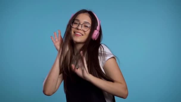 Hezká mladá asijská dívka s dlouhými vlasy baví, usmívá se, tančí se sluchátky ve studiu na modrém pozadí. Hudba, tanec, koncept rádia, zpomalení - Záběry, video