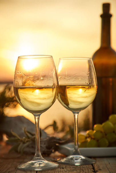 δύο ποτήρια με κρασί και μπουκάλι με σταφύλι κατά τη διάρκεια του ηλιοβασιλέματος - Φωτογραφία, εικόνα