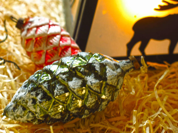 Рождественский фонарь с оленем на фоне опилок с новогодними стеклянными игрушками: зеленый и красный заснеженный конус и гроздь винограда
 - Фото, изображение