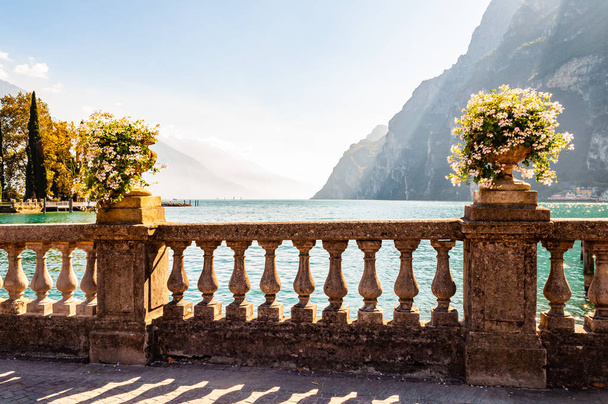 Bellissima passeggiata sul lago di Garda con classiche ringhiere in pietra costruite sul bordo con vasi da fiori con fiori bianchi in fiore. Lago di Garda circondato dalle alte montagne dolomitiche sullo sfondo
 - Foto, immagini