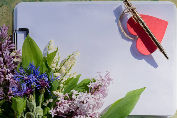 Geburtstag, Mütter, Valentinen, Frauen, Hochzeitstagskonzept. Ein Strauß lila, weißer Frühlingsblumen, ein Notizblock zum Schreiben auf eine Feder und ein fliederfarbener Stift. Platz für Text. Glückwunsch zum Urlaub - Foto, Bild