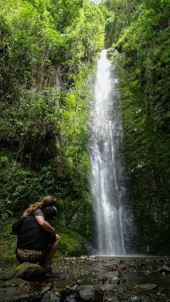 pareja abrazada mirando la caida de una cascada rodeada de vegetaciona Paar umarmt sich, während es einen wunderschönen Wasserfall inmitten der Vegetation beobachtet - Foto, Bild