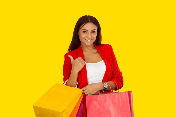 Gyönyörű nő, feltűri a táblát, miközben egy csomó bevásárlószatyrot tart a kezében. Vegyes faji modell elszigetelt sárga háttérrel fénymásoló. Vízszintes kép. Természetes, nincs smink.. - Fotó, kép