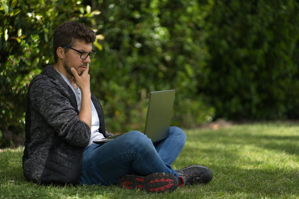 眼鏡をかけた青年がノートパソコンを持って公園の芝生の上に腰を下ろしている。自然環境. - 写真・画像
