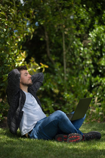 Jovem sentado na grama com seu computador em suas pernas enquanto se estende e relaxa em um parque
 - Foto, Imagem