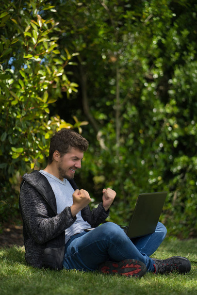 Young man sitting on a park while watching his computer and celebrating furiously with his hands. Natural Environment. - Foto, Imagem
