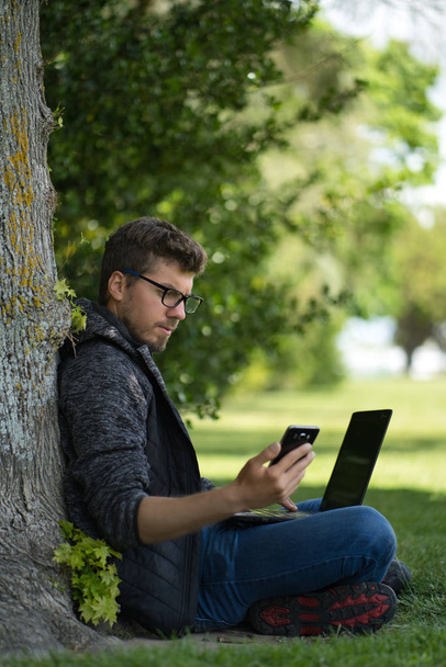 Bilgisayarında çalışan genç beyaz bir adam aynı anda telefonuna bakarken bir ağacın altında oturuyor. Teknolojik konsept - Fotoğraf, Görsel