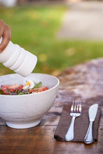 Mão com uma saladeira de pimenta branca temperando uma tigela de salada fresca folhas verdes misturadas, ovos, azeitonas pretas e tomate em uma mesa de madeira com talheres
 - Foto, Imagem