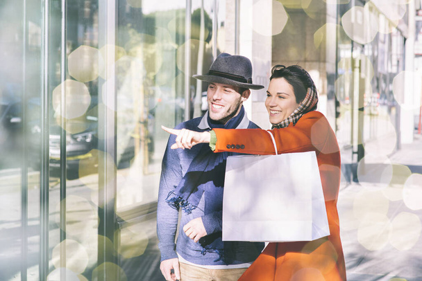 ホリデーシーズン中に冬服のウィンドウショップで身に着けている若い男と女性-笑顔でポインティングと幸せ-ボケ照明と日中の街で - 写真・画像