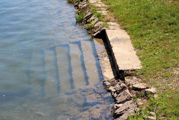 Passos de concreto inundado com rio lamacento na margem do rio local cercado com pedras e grama não cortada no dia ensolarado quente do verão
 - Foto, Imagem
