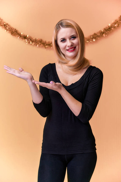 Portrait of a cute girl in a black T-shirt with long beautiful hair and great makeup. Concept of a young blonde woman with New Year's decor. Smiling, showing emotions on a pink background. - Photo, Image
