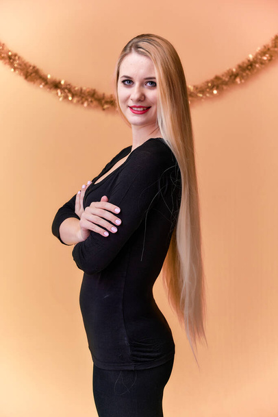 Portrait of a cute girl in a black T-shirt with long beautiful hair and great makeup. Concept of a young blonde woman with New Year's decor. Smiling, showing emotions on a pink background. - Photo, image