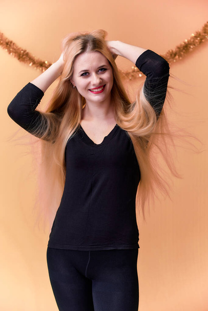 Portrait of a cute girl in a black T-shirt with long beautiful hair and great makeup. Concept of a young blonde woman with New Year's decor. Smiling, showing emotions on a pink background. - Fotó, kép