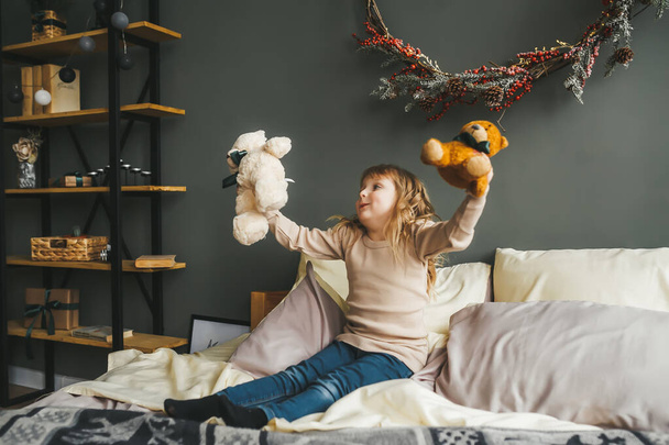 Menina bonita brincando com um brinquedo de ursinho sentado em uma cama perto de uma árvore de natal
. - Foto, Imagem
