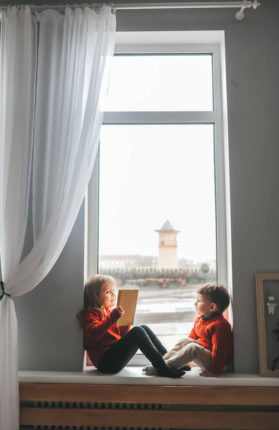 A fiú és a nővére otthon játszanak az ablakpárkányon és könyveket olvasnak.. - Fotó, kép