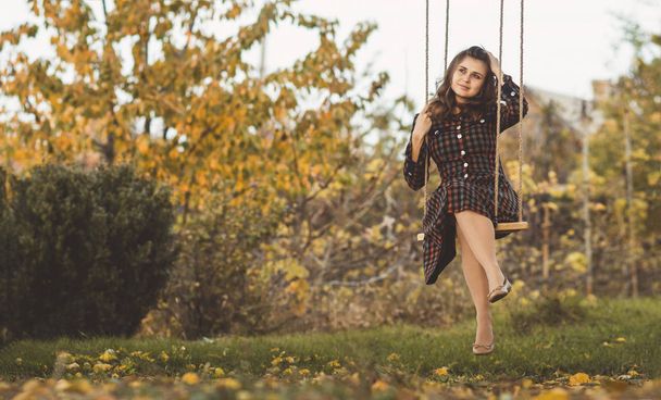 cute girl in a dress sways on a swing in the autumn garden, a young woman in a positive romance mood relaxing on nature - Photo, Image