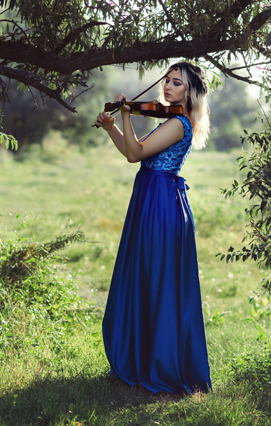 beautiful girl plating violin under tree outdoors, young woman in long elegant dress with a musical instrument on nature make performance, concept music and art - Photo, Image