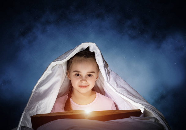 Happy little girl reading interesting book in bed after bedtime. Beautiful child with flashlight hiding under blanket. Smiling girl in pajamas on deep blue sky background. Child reading fairy tales. - Fotó, kép