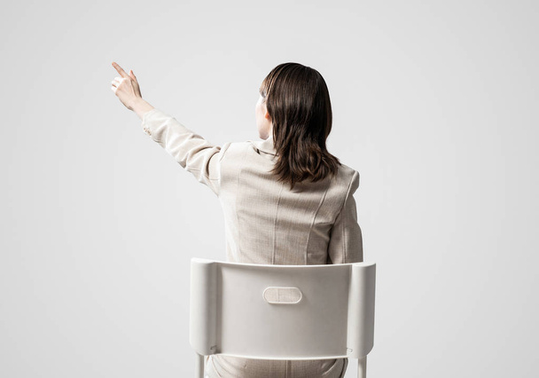 Woman presenting or something finger pointing. Back view long haired woman in white suit sitting on white chair. Business concept with copy space. Template for advertising or business presentation - 写真・画像