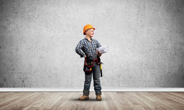 Attractive female architect in hardhat standing with technical blueprints. Portrait of young worker in checkered blue shirt and jeans standing in empty room. Interior design and renovation service. - Φωτογραφία, εικόνα