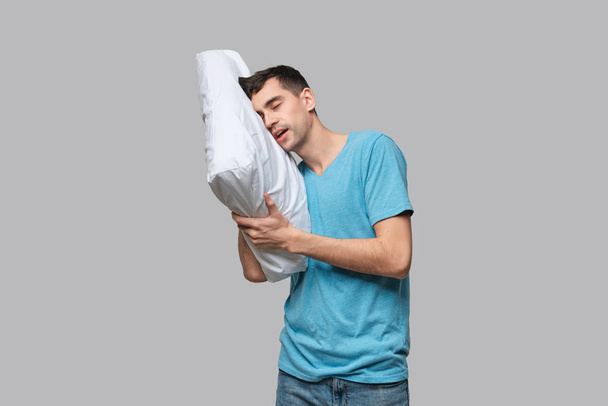 Tired brunet man in a blue tee resting on a white pillow isolated over grey background. - Photo, Image