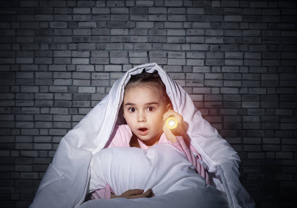 Frightened girl with flashlight hiding under blanket. Scared kid lying in his bed at home. Night terrors of child. Little girl can not sleep at night. Child in pajamas on background of grey wall. - Photo, Image
