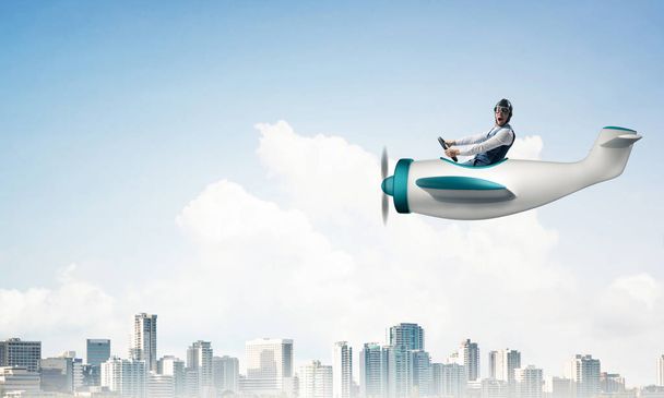 高い近代的な建物とダウンタウンの上にプロペラ飛行機を駆動飛行士の帽子とゴーグルのビジネスマン。小さな飛行機で楽しんでいる面白い男。雲の背景を持つ青い空 - 写真・画像